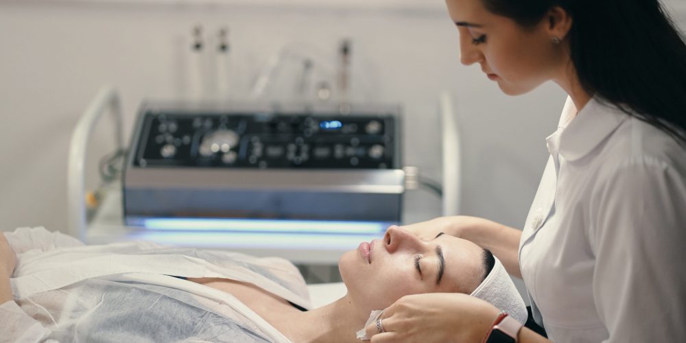 Woman lying during cosmetic procedure while skillful female cosmetologist making facial treatment